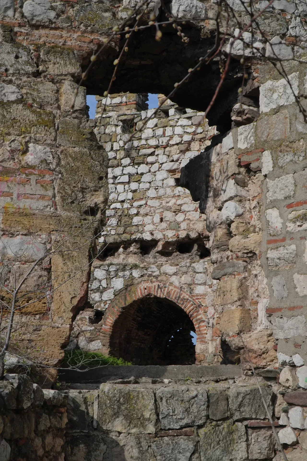 Drevni kameni kompleks sa zaobljenim prolazom i otvorima u zidovima kroz koje se vidi plavo nebo. Gole grane sa pupoljcima se protežu odozgo u kadar.