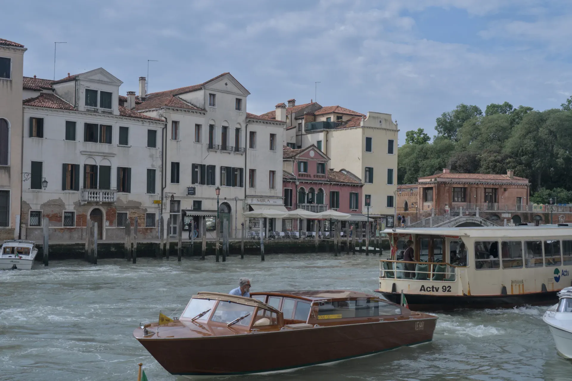 Un hombre con una camisa azul claro pilota un taxi acuático de madera clásico en un canal de Venecia, Italia, con otros barcos y edificios históricos visibles al fondo bajo un cielo nublado.