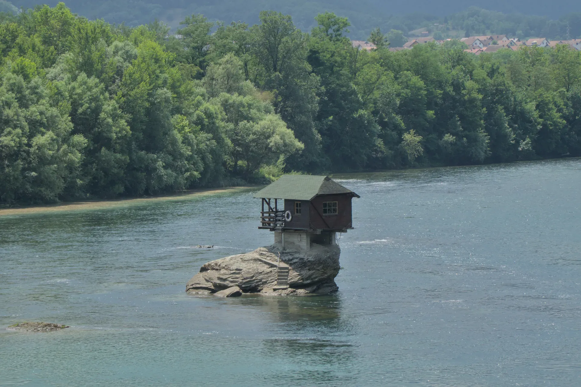 A small, wooden house with a green roof is situated on a rock in the middle of the Drina River. Steps lead down to the water from the rock. Lush green trees line the riverbank in the background, within Tara National Park, Serbia. The house is known as 'домик на Дрине'.