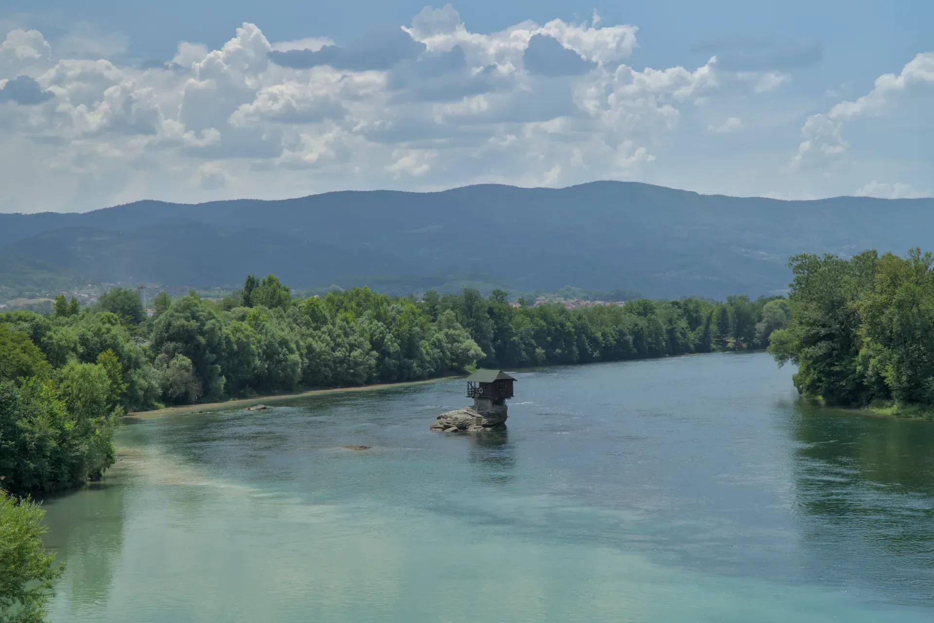 Una pequeña casa posada sobre una roca en el río Drina en Serbia, con colinas verdes y orillas boscosas al fondo.