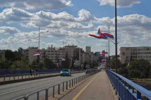 A bridge in Novi Sad, Serbia, with cars and pedestrians, decorated with numerous Serbian flags fluttering in the wind against a backdrop of buildings and clouds.