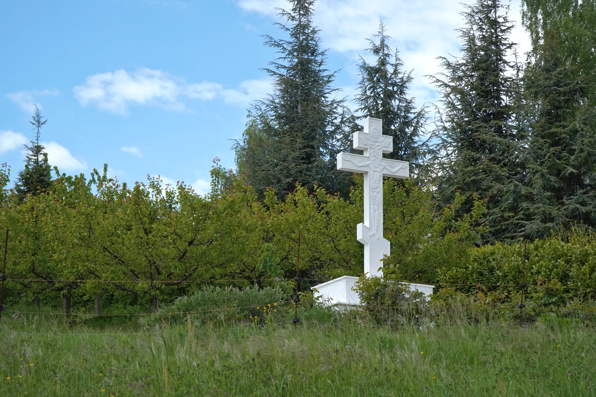 A white Orthodox cross with intricate carvings stands tall on Fruska Gora mountain, near Stari Hopovo, set against a backdrop of dense green trees and a blue sky with scattered clouds, in Novi Sad, Serbia.