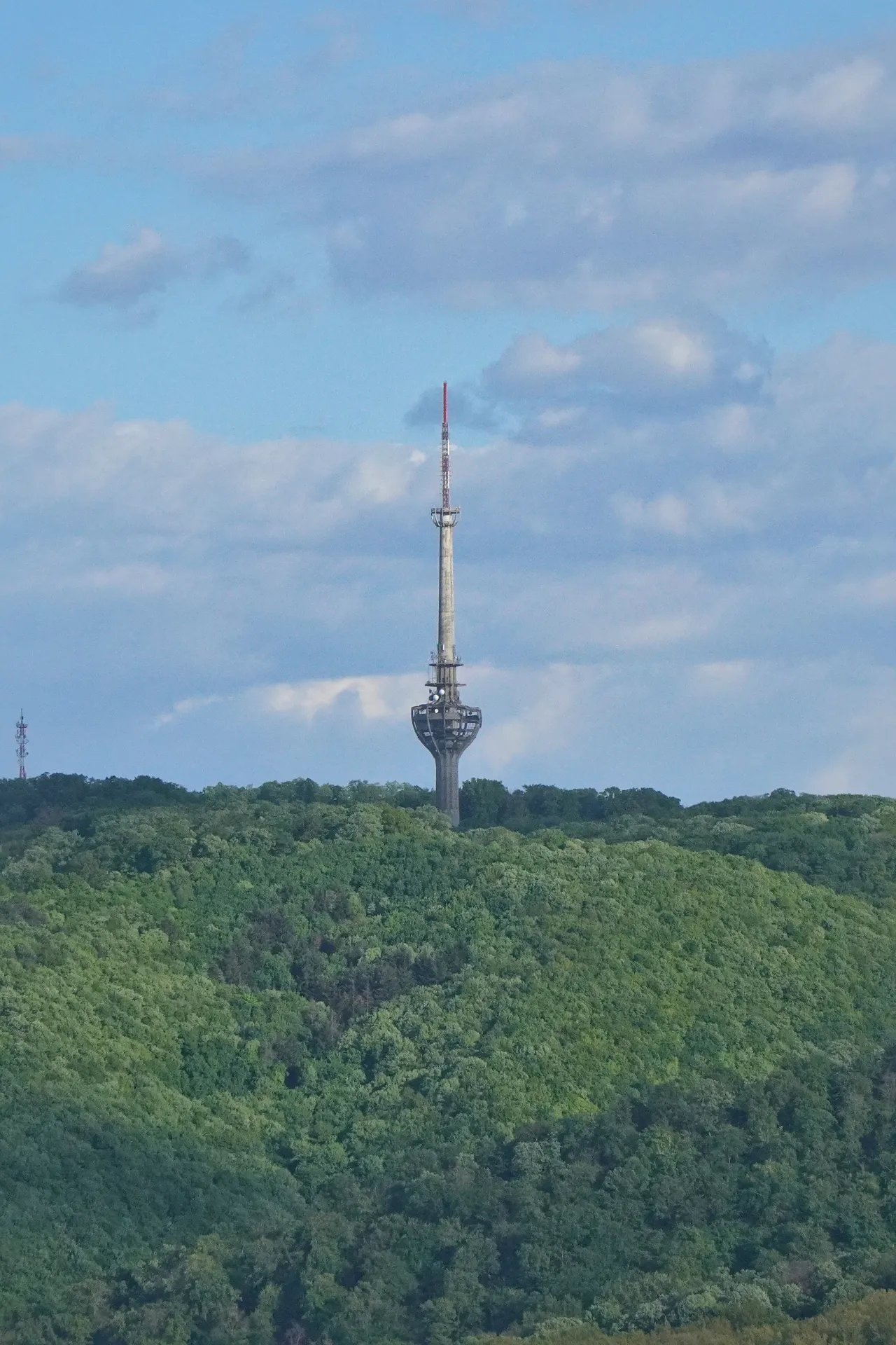 The telecommunication tower on Fruška Gora, Serbia, with lush green trees below and a blue sky with clouds above.