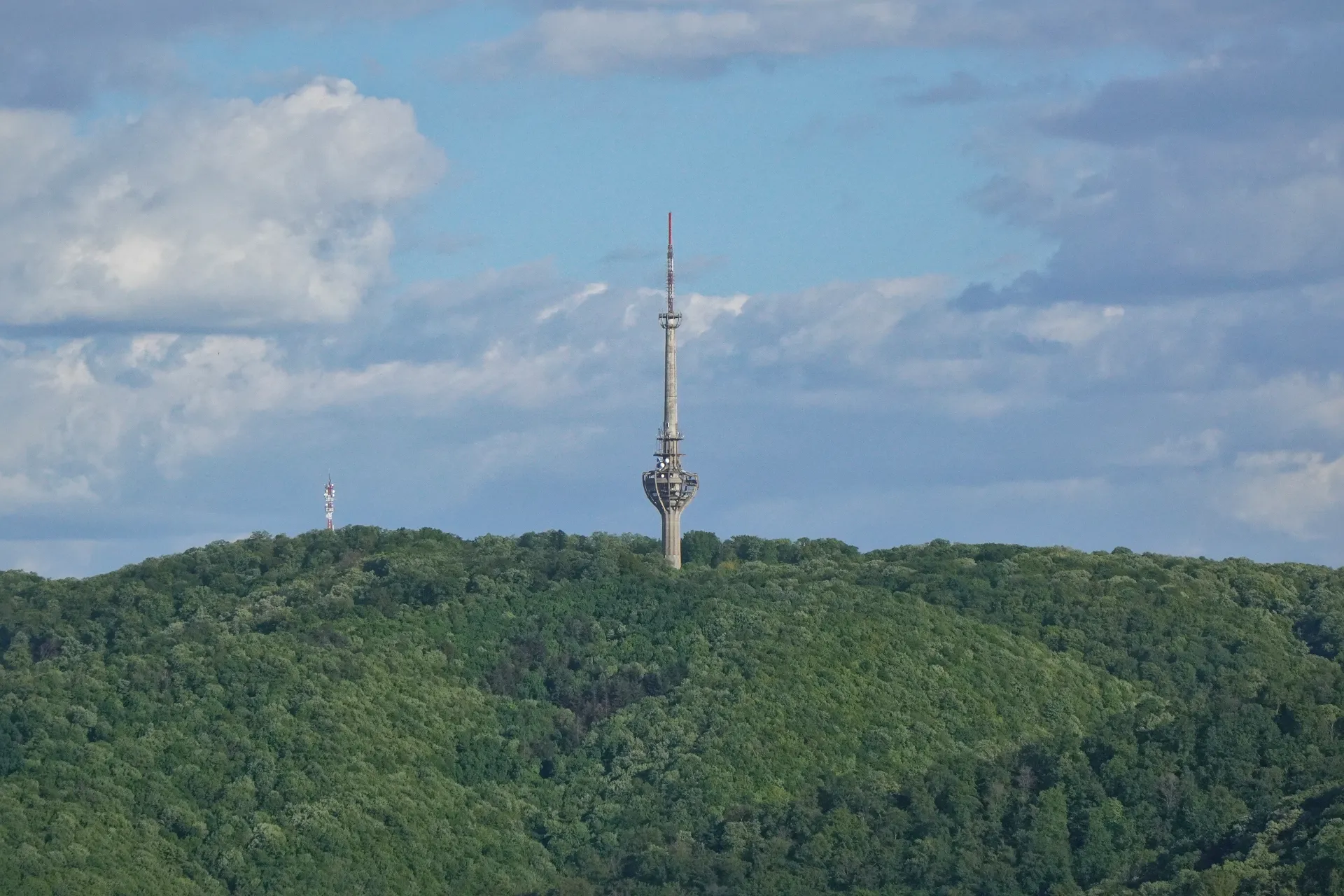 The television tower on Fruška Gora, Serbia, rises above a dense green forest under a cloudy blue sky.