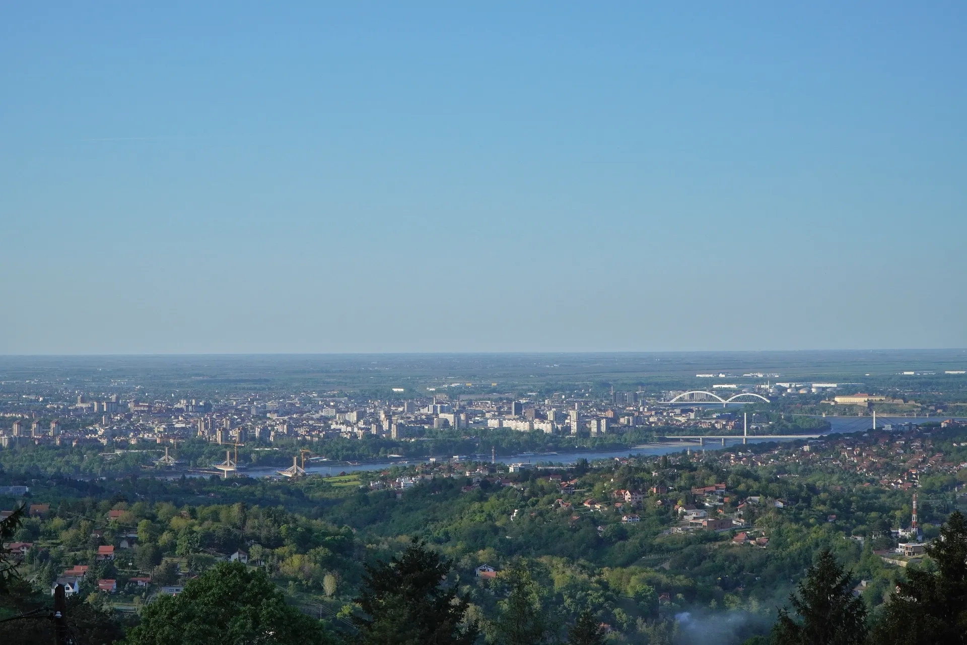 Panoramski pogled na Novi Sad, Srbija, koji prikazuje gradski horizont, reku Dunav sa mostovima i zelena brda Fruške Gore u prvom planu pod vedrim plavim nebom.