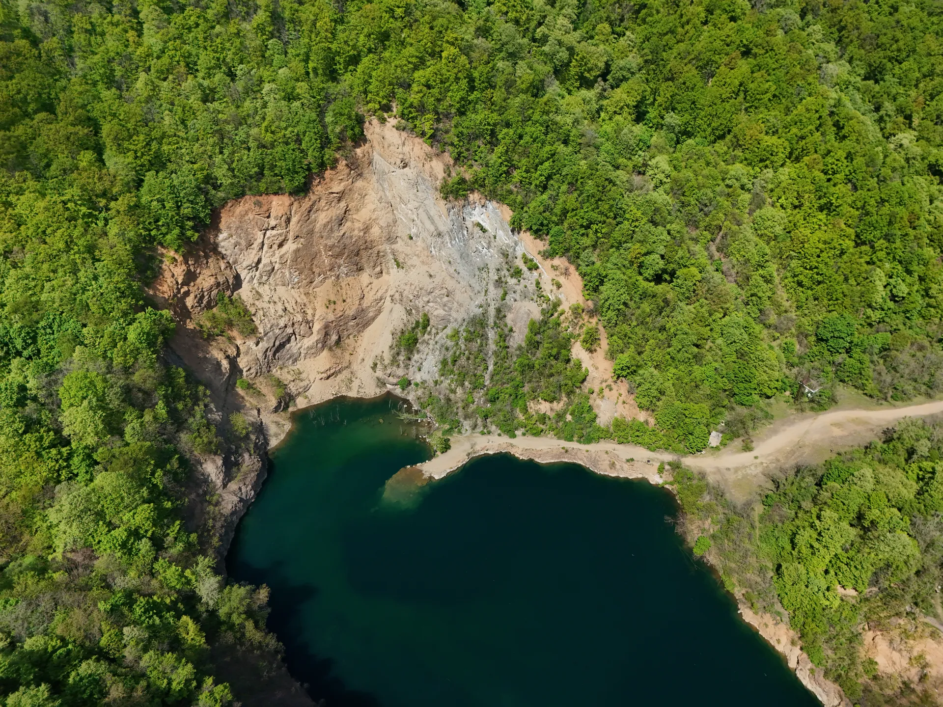 Aerial view of Ledinačko Jezero (Ledinci Lake) near Novi Sad, Serbia, showcasing the deep blue water surrounded by dense green forests and a rocky cliff.