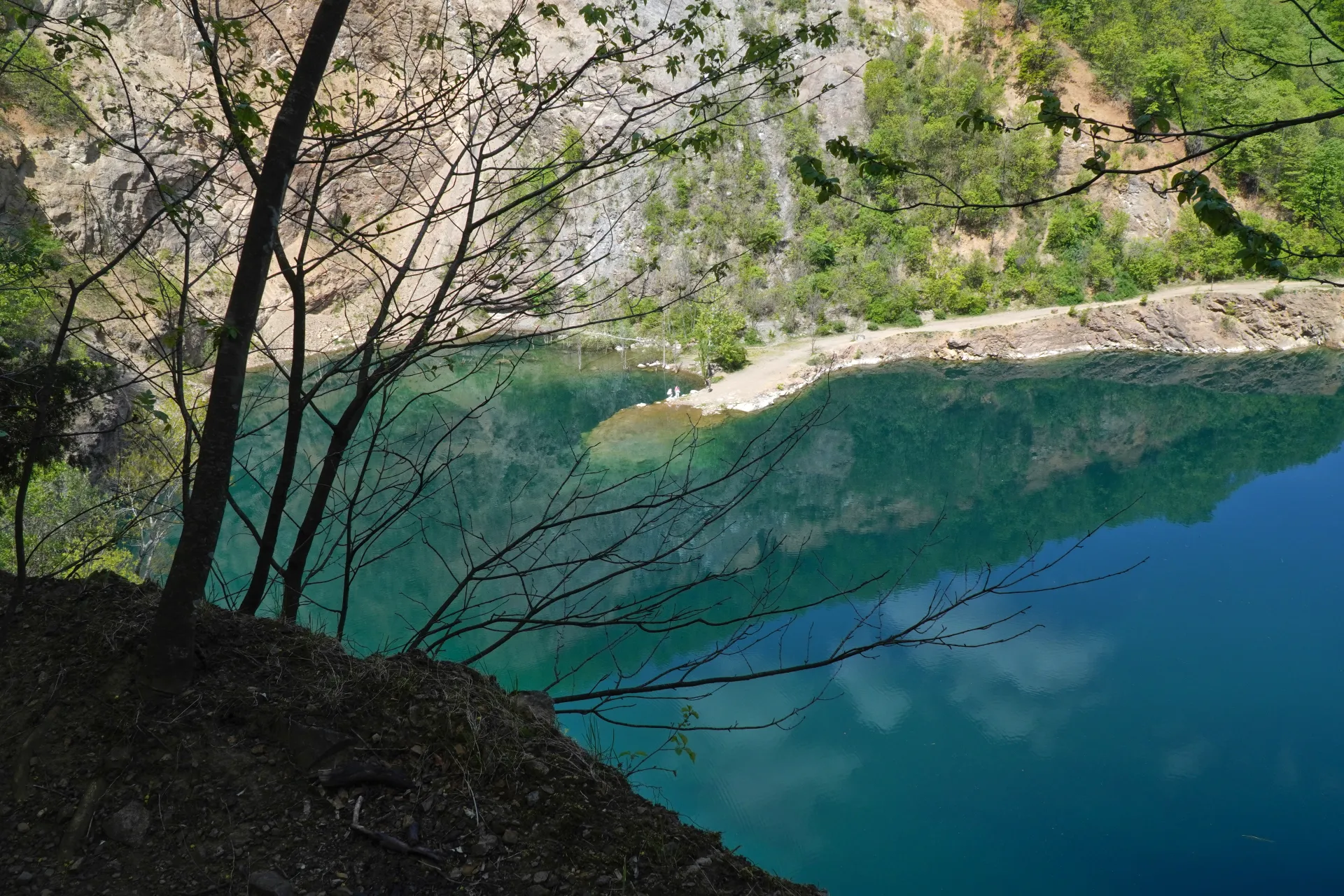 View of Ledinačko Lake with its clear turquoise water and surrounding rocky cliffs, framed by bare tree branches.