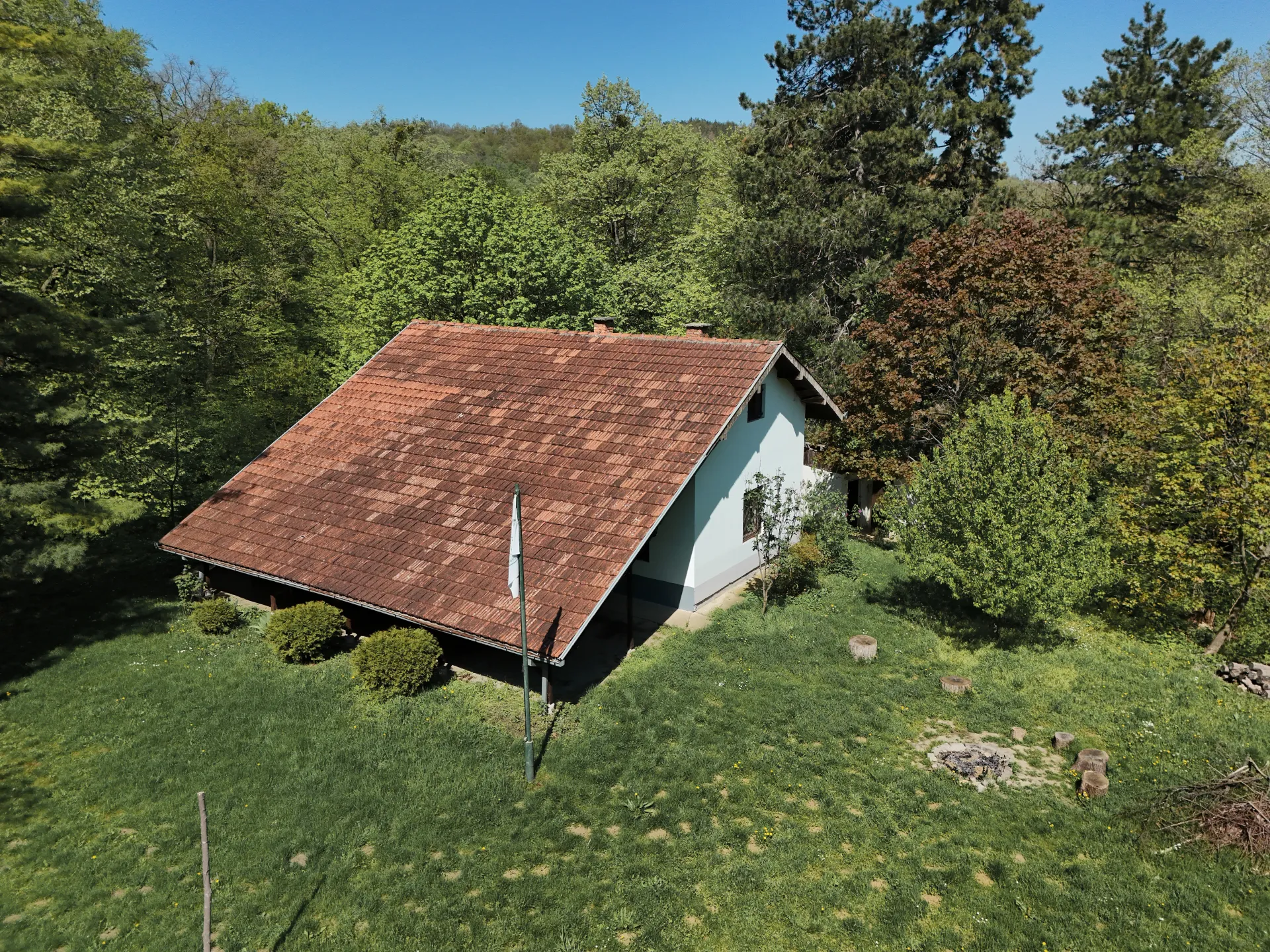 A cozy house with a tiled roof sits in a green meadow, surrounded by trees. A flag is planted in the yard, and a fire pit with log seats is visible nearby, indicating a picnic area and recreation base.