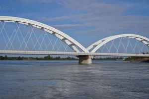 A modern white arch bridge with intricate cable supports spans the Danube River in Novi Sad, Serbia, under a cloudy blue sky.