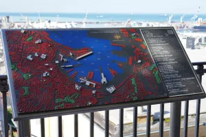 A detailed 3D model map of Genoa, Italy, displaying the city's architecture and port area, with the Ligurian Sea and port cranes visible in the background.