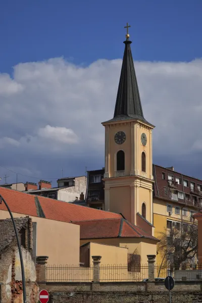 La iglesia en Pančevo, un edificio llamativo con un campanario alto y esbelto coronado por una cruz. La torre tiene una esfera de reloj visible. Junto a la iglesia hay varios edificios urbanos con fachadas rojas y amarillas, todo bajo un cielo nublado dramático.
