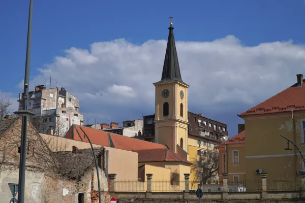 A yellow church tower with a black pointed roof and a clock is visible in Pančevo, Serbia. Modern buildings are in the background, and the sky is blue with white clouds.