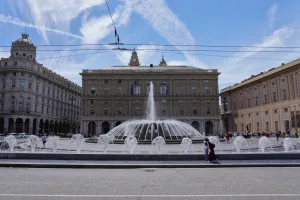 A lively fountain with multiple streams of water jets in action, situated in the main square of Genoa, Italy. Ornate buildings and a clock tower are visible behind the fountain under a bright, cloudy sky.