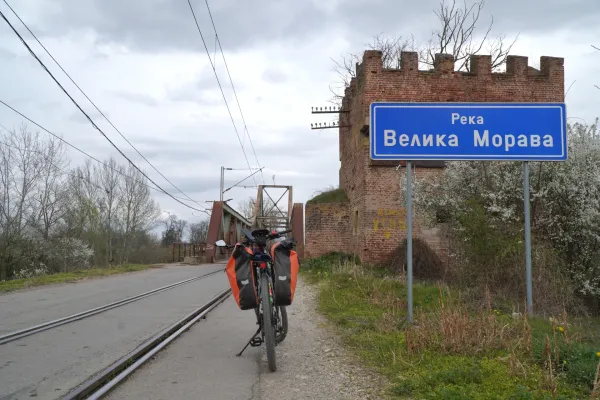 Una bicicletta carica di borse da sella arancioni si trova su un sentiero sterrato accanto ai binari del treno. Sullo sfondo è visibile il Ponte Ljubičevski sul Grande Morava, insieme a un'antica torre di mattoni e a un cartello blu con scritto 'Velika Morava'. La scena si trova vicino Požarevac, Serbia.