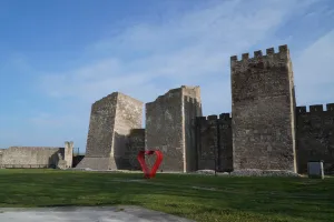 La Fortezza di Smederevo in Serbia con le sue alte mura di pietra e torri, accanto una scultura a forma di cuore rosso brillante su un prato verde.