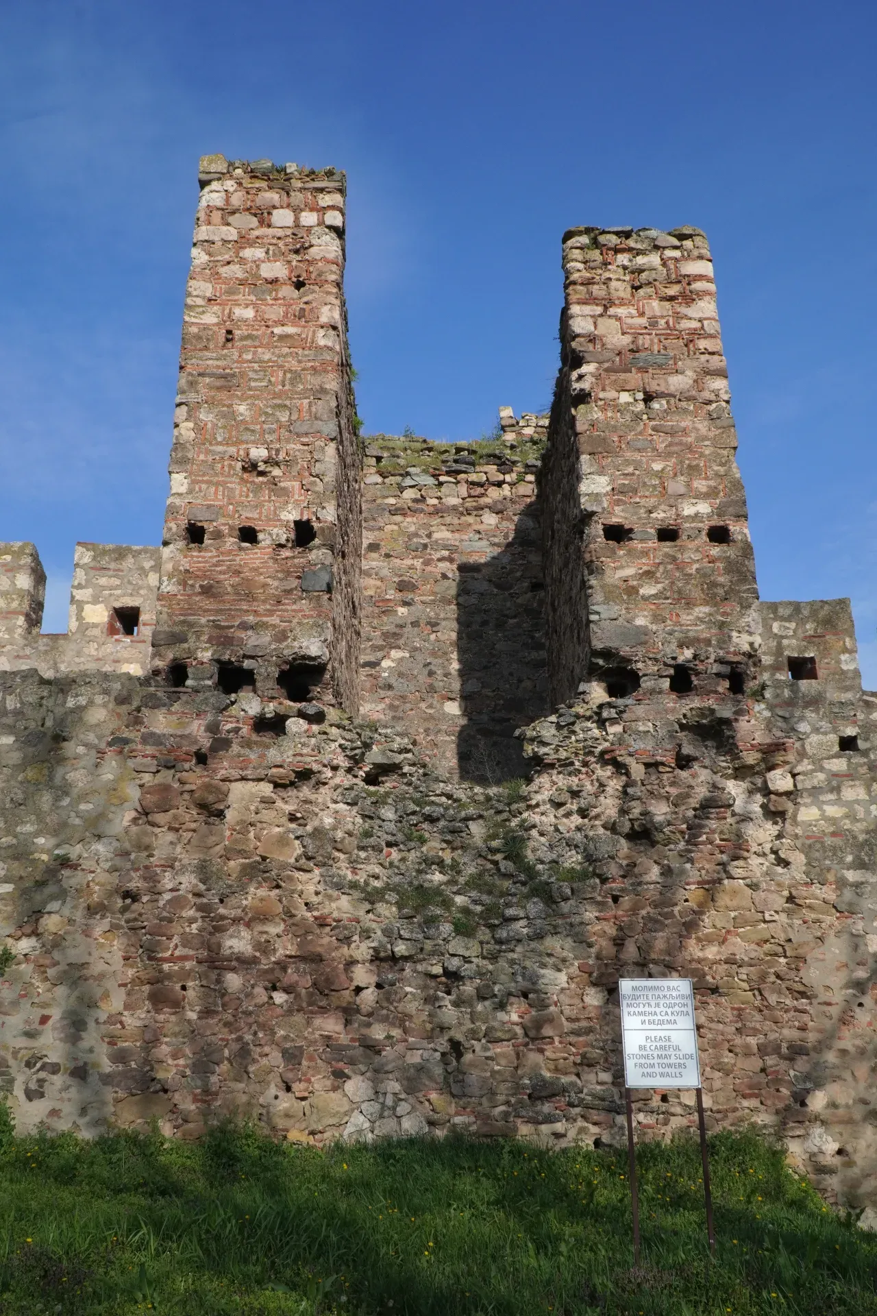Two tall, dilapidated stone towers of the Smederevo Fortress in Serbia, with smaller wall remnants, under a blue sky, with green grass in the foreground.