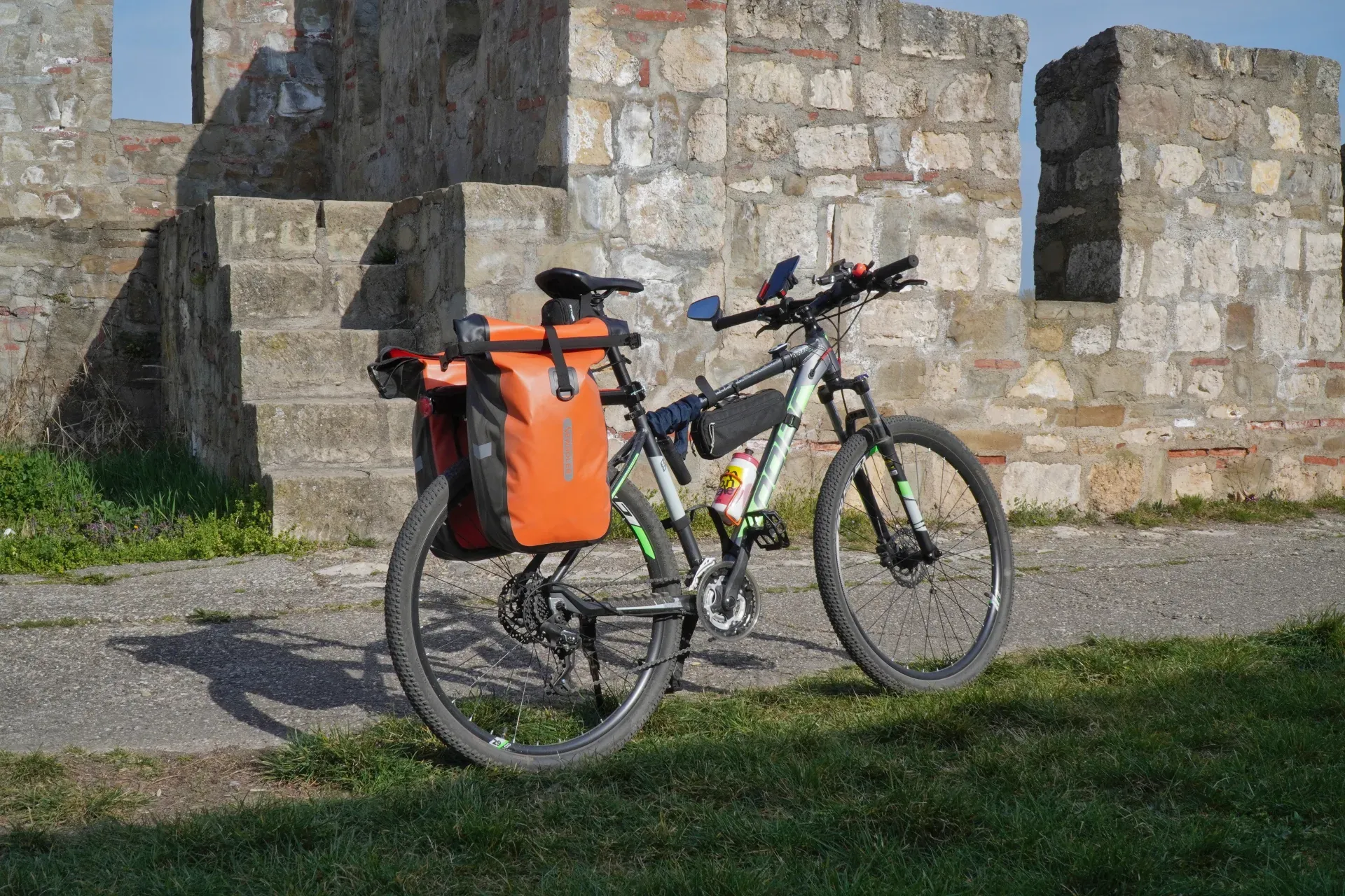 A bicycle loaded with orange panniers stands on a paved path in front of an old stone wall of Smederevo Fortress, Serbia.
