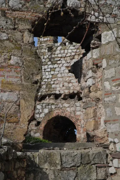 An old stone complex with a rounded passageway and holes in the walls, through which the blue sky can be seen. Some branches with buds protrude from the top into the image.