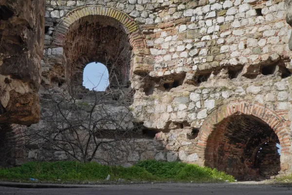 Un muro di pietra fatiscente con diverse arcate e aperture attraverso cui si vede il cielo azzurro, così come rami secchi e vegetazione verde a terra.