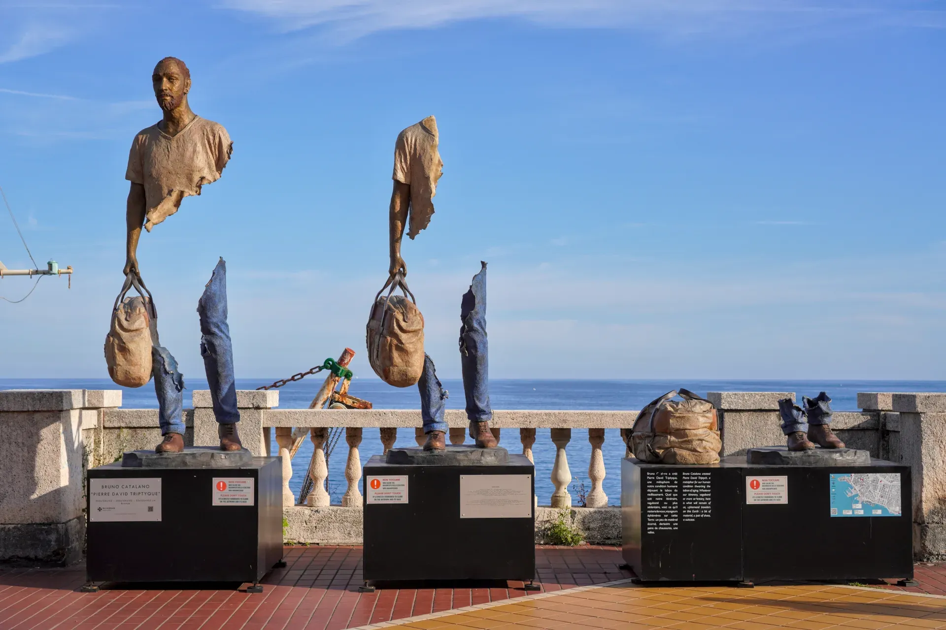 Fragmented bronze sculptures of human torsos and legs carrying bags, displayed on plinths along the Genoa waterfront with the blue sea and sky in the background.