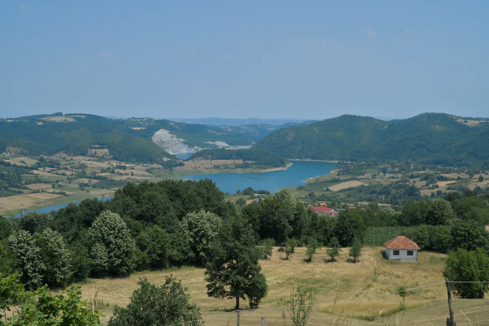 A vast landscape with green, forested hills, cultivated fields, and a turquoise lake in Tara National Park, Serbia. A small white house with a red roof stands in a field surrounded by trees. In the background, more hills and the wide sky are visible.