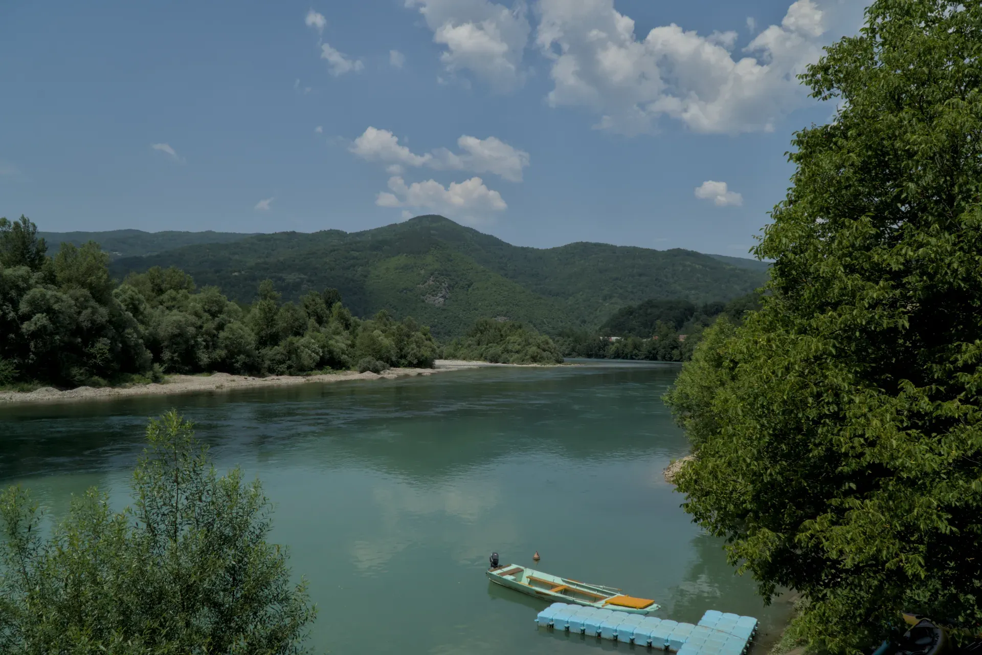 A wide view of the Drina river with green, forested hills in the background. A small boat is moored on the bank with a floating dock beside it. The turquoise water is calm and reflects the blue sky with scattered clouds.