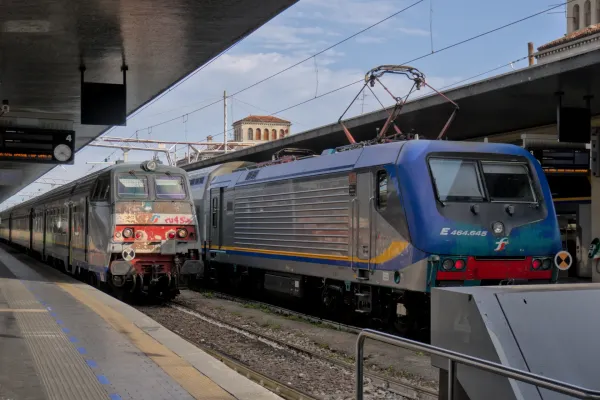 Zwei Züge auf Gleisen in einem italienischen Bahnhof mit historischer Architektur im Hintergrund.