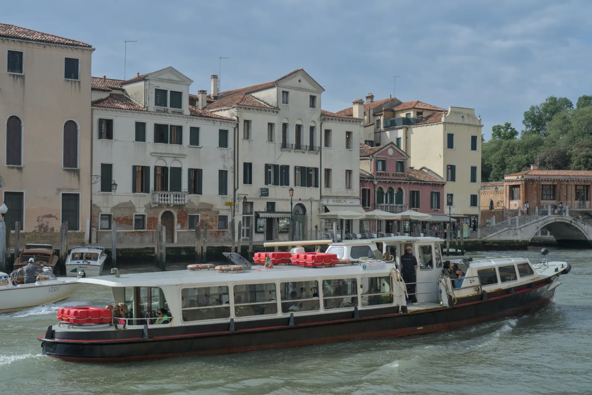 A vaporetto sails on a canal in Venice, surrounded by historic buildings and a pedestrian bridge in the background.