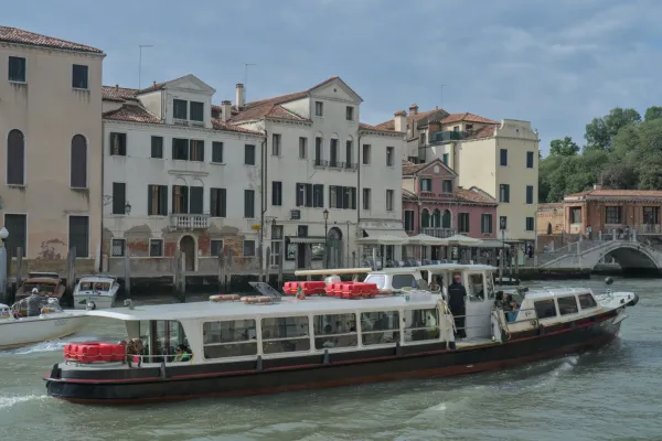 Ein Vaporetto fährt auf einem Kanal in Venedig, umgeben von historischen Gebäuden und einer Fußgängerbrücke im Hintergrund.
