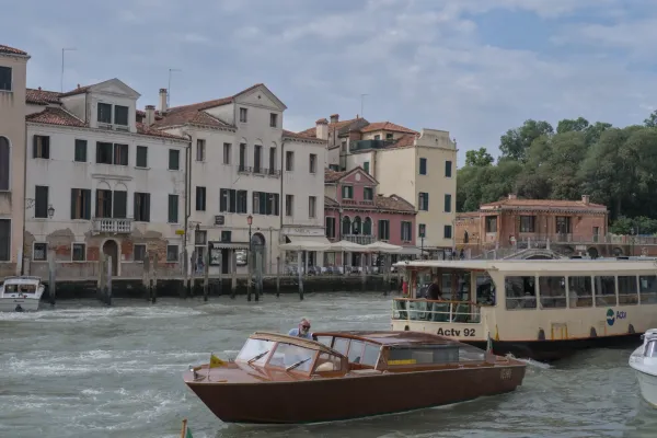 Venecijanski kanal sa prednjim pogledom na braon taksi čamac i veći putnički brod, okružen klasičnim zgradama pod oblačnim nebom.