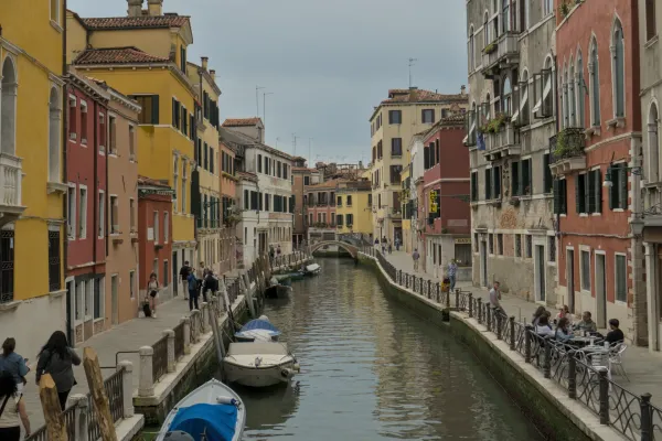 Uzak prolaz u Veneciji sa šarenim kućama, kanalom i čamcima, sa ljudima koji šetaju trotoarom.