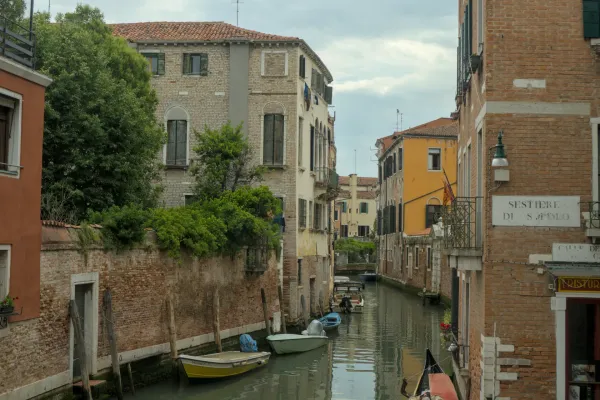 Uzak venecijanski kanal sa tradicionalnim zgradama, zelenilom i nekoliko čamaca koji plutaju u vodi.