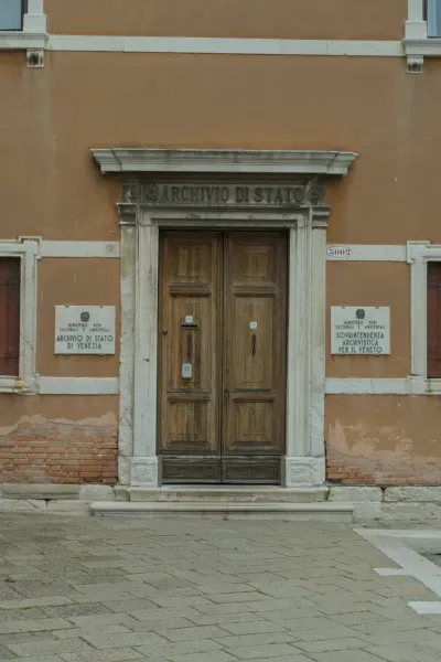 Široka, stara drvena vrata na ulazu u Archivio di Stato u Veneciji, uokvirena kamenim stubovima i oker fasadom.