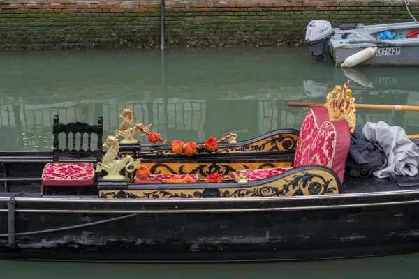 Raskošna mletačka gondola sa crvenim i zlatnim ukrasima, koja pluta na mirnoj zelenoj vodi pored ciglanog zida.