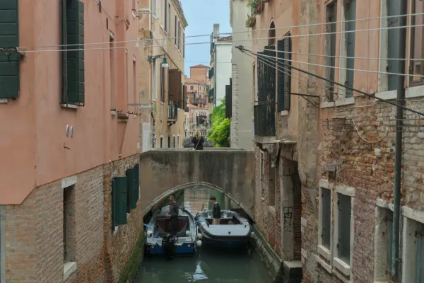Venecijanski kanal sa dva čamca i ljudima ispod mosta, okružen starim zgradama od cigle.