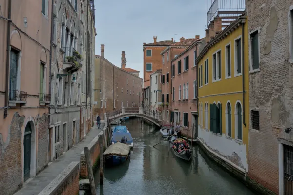 Uski kanal u Veneciji sa gondolom, duž kojeg se nižu šarene istorijske zgrade i šetalište.