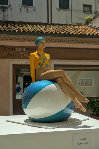 Porträt einer Skulptur einer Frau, die auf einem blauen und weißen Strandball in Venedig sitzt und eine gelbe Badehose trägt.