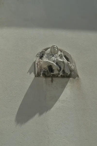Skulptur eines Engels mit Flügeln an einer strukturierten, hellen Wand mit einem dramatischen Schattenwurf.