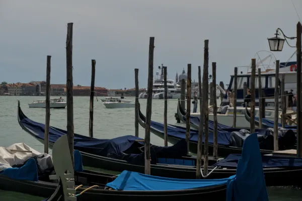 Niz venecijanskih gondola, sa plavim pokrivačima, na pozadini grada i vode, sa drugim čamcima u pozadini.