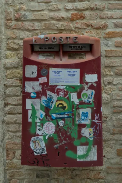 Stari crveni poštanski sanduče ukrašen nalepnicama i grafitima ispred ciglanog zida u Veneciji.