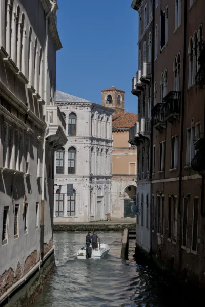 Čamac plovi uskim venecijanskim kanalom, okružen istorijskim zgradama, sa zvonikom u pozadini.