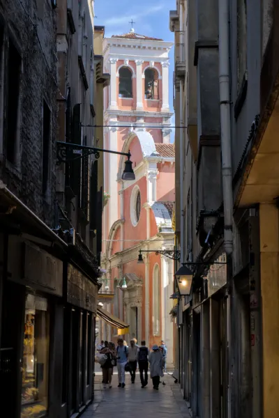 Fotografija uske mletačke ulice koja vodi ka roze zvoniku sa zvonima i lukovima, sa ljudima koji prolaze.