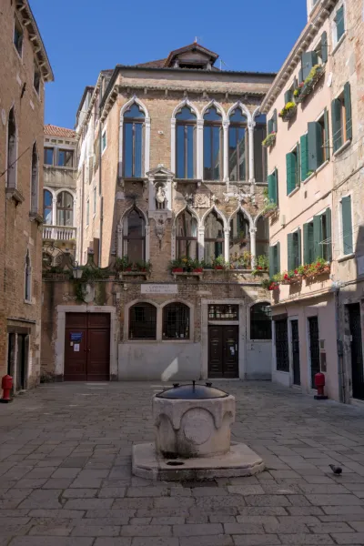 Ein sonniger venezianischer Innenhof mit einem steinernen Brunnen im Vordergrund und historischen Gebäuden mit Bögen.