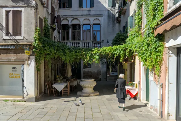 Žena prolazi kroz zelenu ulicu u Veneciji obavijenu bršljanom, sa stolovima na otvorenom i starinskim bunarom.