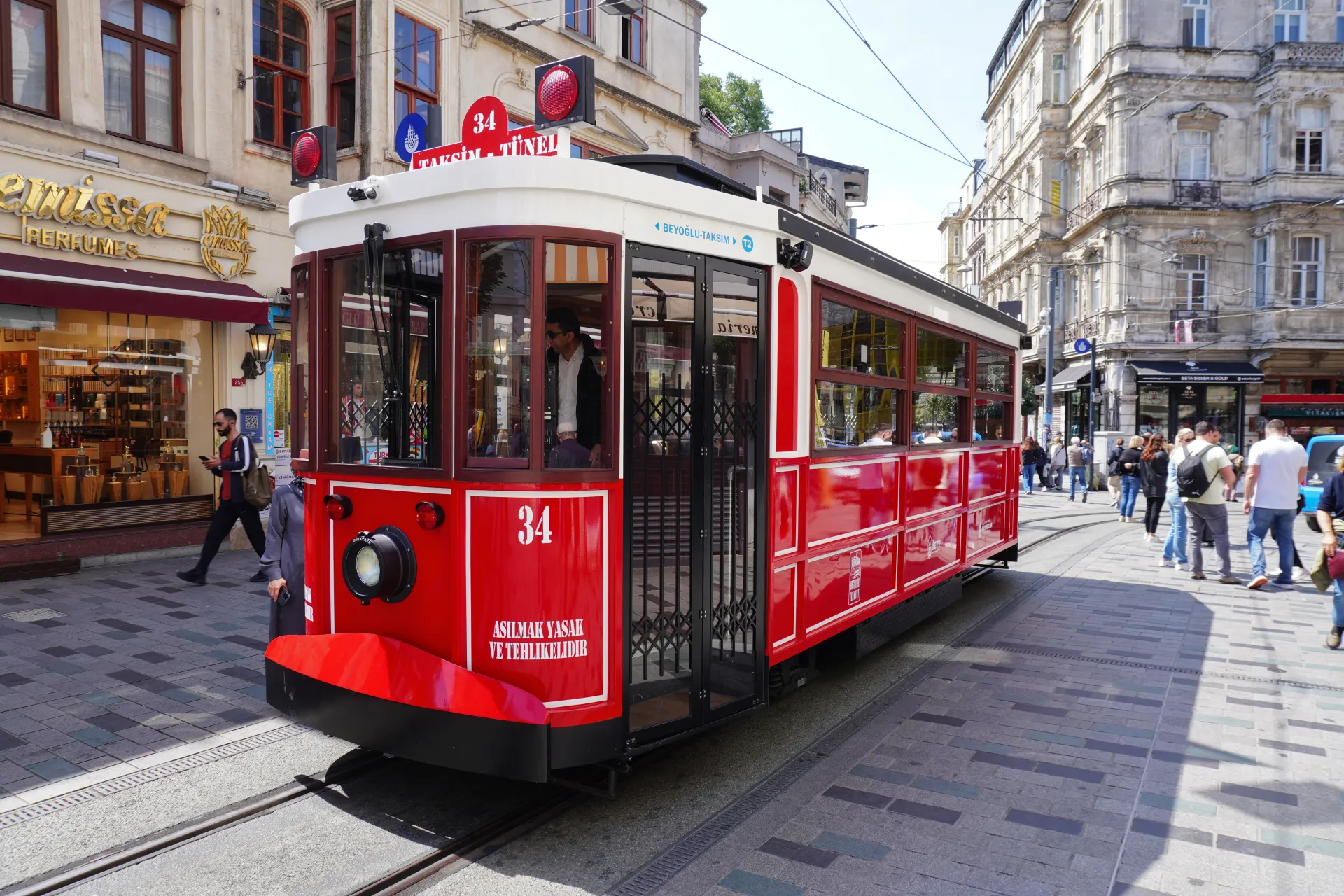 A bright red tram with route number 34 and the destination 'Beyoglu-Taksim' travels down a cobblestone street in Istanbul, Turkey, past shops and pedestrians.