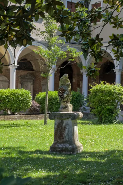 Eine steinerne Skulptur auf einem Sockel, umgeben von einem üppigen grünen Garten auf der Friedhofsinsel San Michele in Venedig.