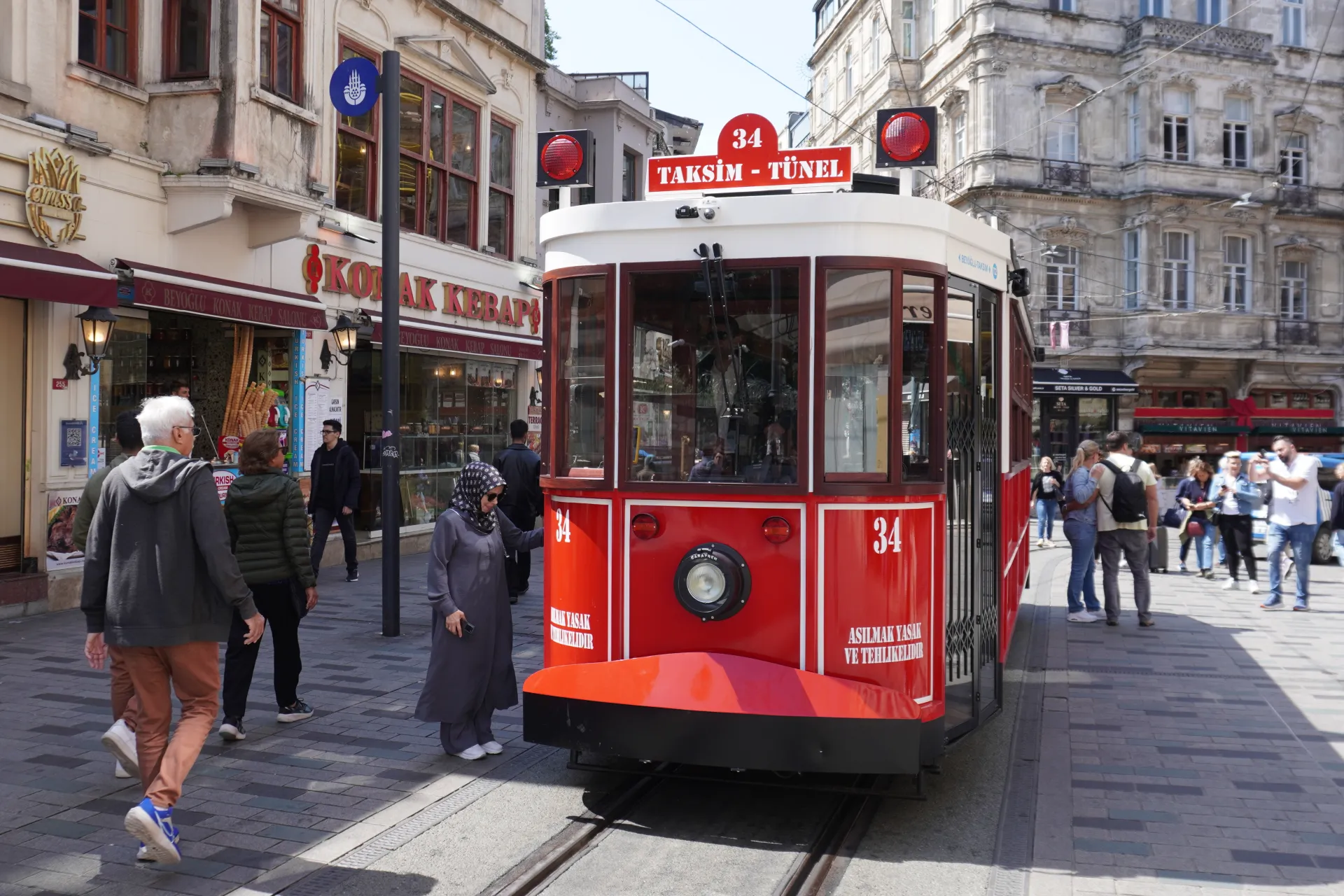 A vintage red tram with the route '34 Taksim - Tünel' marked on it, standing on tram tracks on a pedestrian street in Istanbul. People are walking on the sidewalk next to the tram and in front of shops with signs like 'KOTAK KEBAP'.