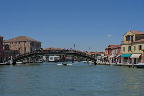 Drveni pešački most na ostrvu Murano u Veneciji, preko koga prelaze ljudi, sa čamcima na vodi i istorijskim zgradama.
