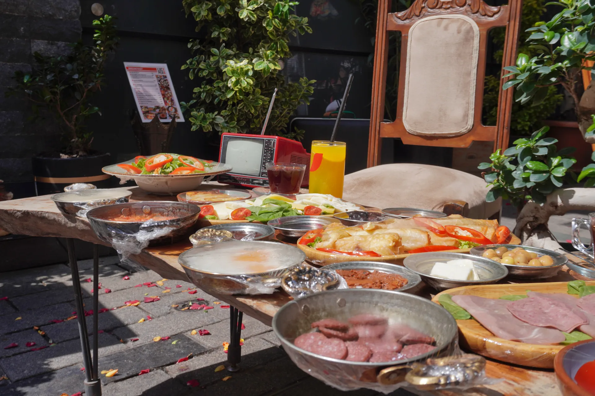 A lavish Turkish breakfast spread featuring various dishes like sucuk, cheese, olives, and salads on a wooden table, with a vintage TV and drinks in the background.