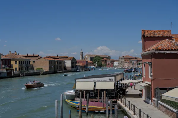 Pogled na obalu kanala ostrva Murano sa vodom, čamcima i karakterističnim venecijanskim kućama pod plavim nebom.